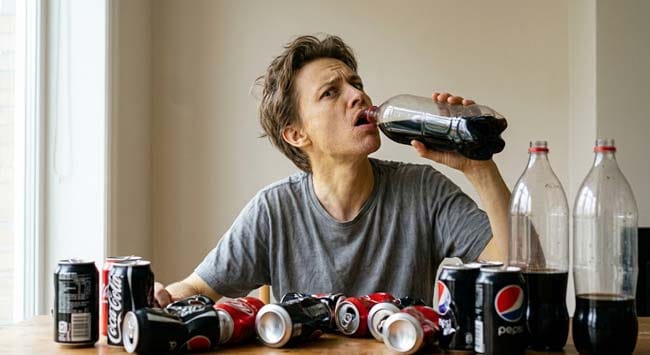 A person drinking cola surrounded by empty cola bottles with no water visible illustrating what happens when cola replaces water causing osmotic dehydration phosphoric acid calcium loss and fructose liver overload