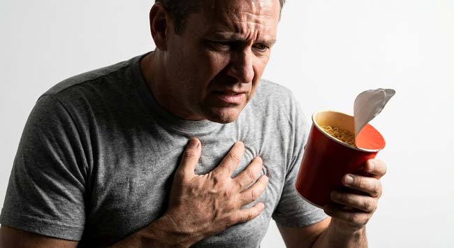 A person clutching their chest in cardiac discomfort while holding instant noodles illustrating the four cardiovascular mechanisms of instant noodles effects on heart including sodium hypertension arterial plaque TBHQ calcification and potassium arrhythmia risk