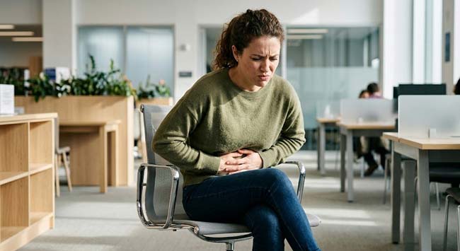 A person looking visibly uncomfortable holding their stomach and crossing their legs illustrating what happens if you hold your poop and the progressive colon water absorption that hardens stool into stone-like masses