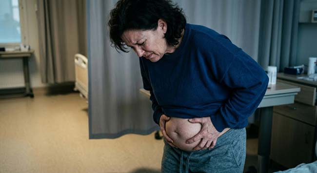 A person clutching their lower abdomen in severe pain with visible distension illustrating the symptoms of fecaloma where chronic stool retention creates a mineralized stone-like mass requiring medical removal